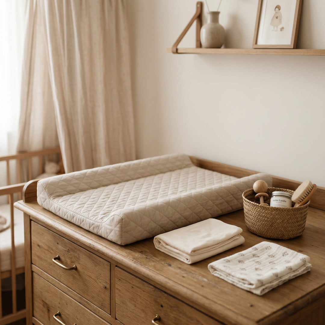 Matelas à langer matelassé beige à rebords posé sur une commode en bois, avec langes pliés et panier d'accessoires de soin